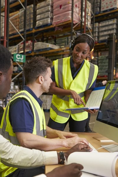 In a warehouse, a supervisor in a bright yellow vest points to a clipboard and speaks with her coworkers also wearing yellow vests