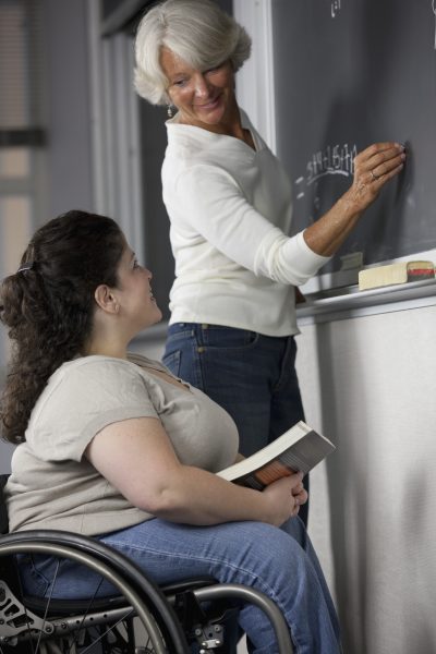 A teacher writes on a chalkboard while looking at her student, who is in a wheelchair