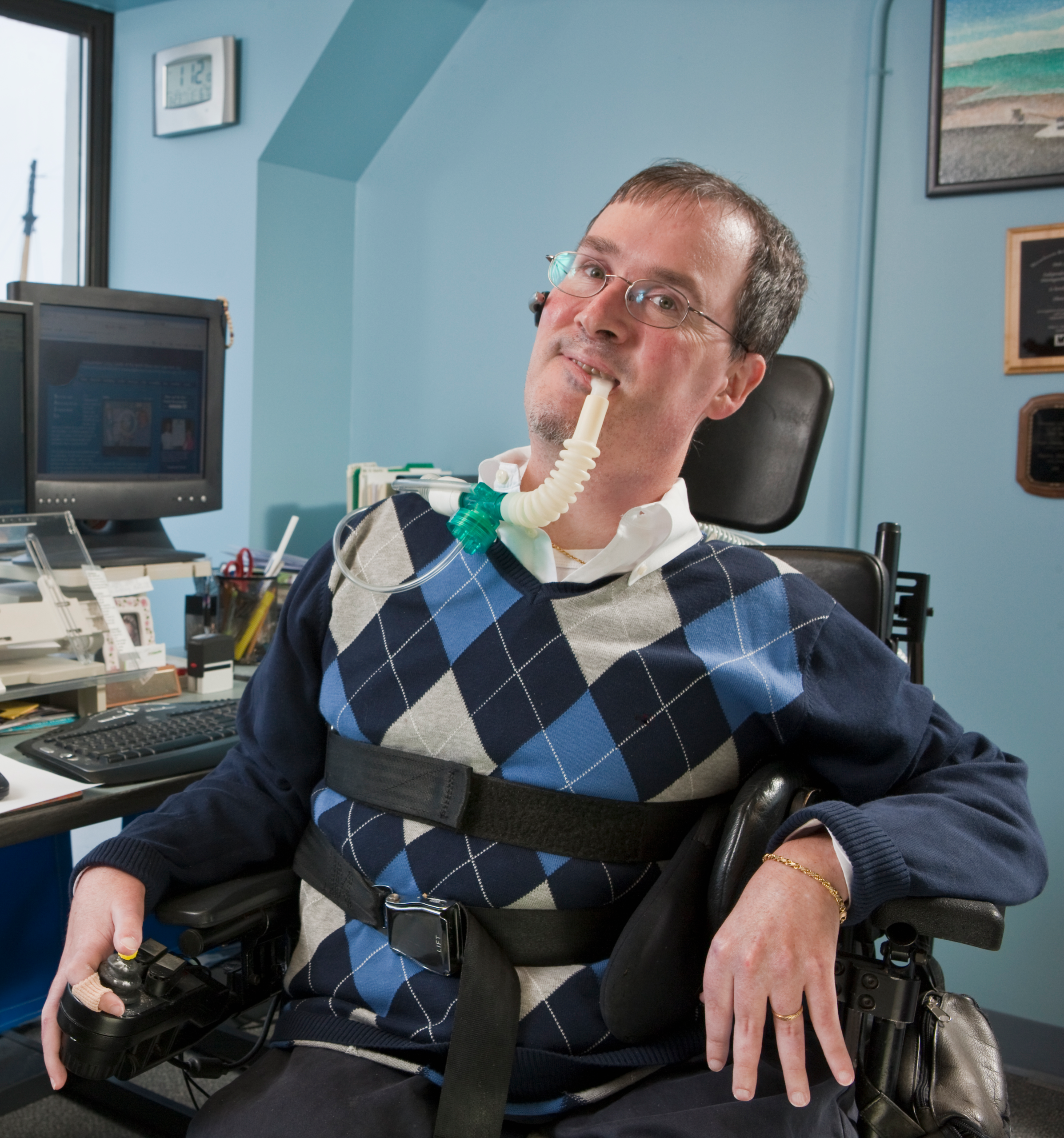 A man with a disability strapped into a wheelchair sits in front of a computer. He also has a feeding tube.