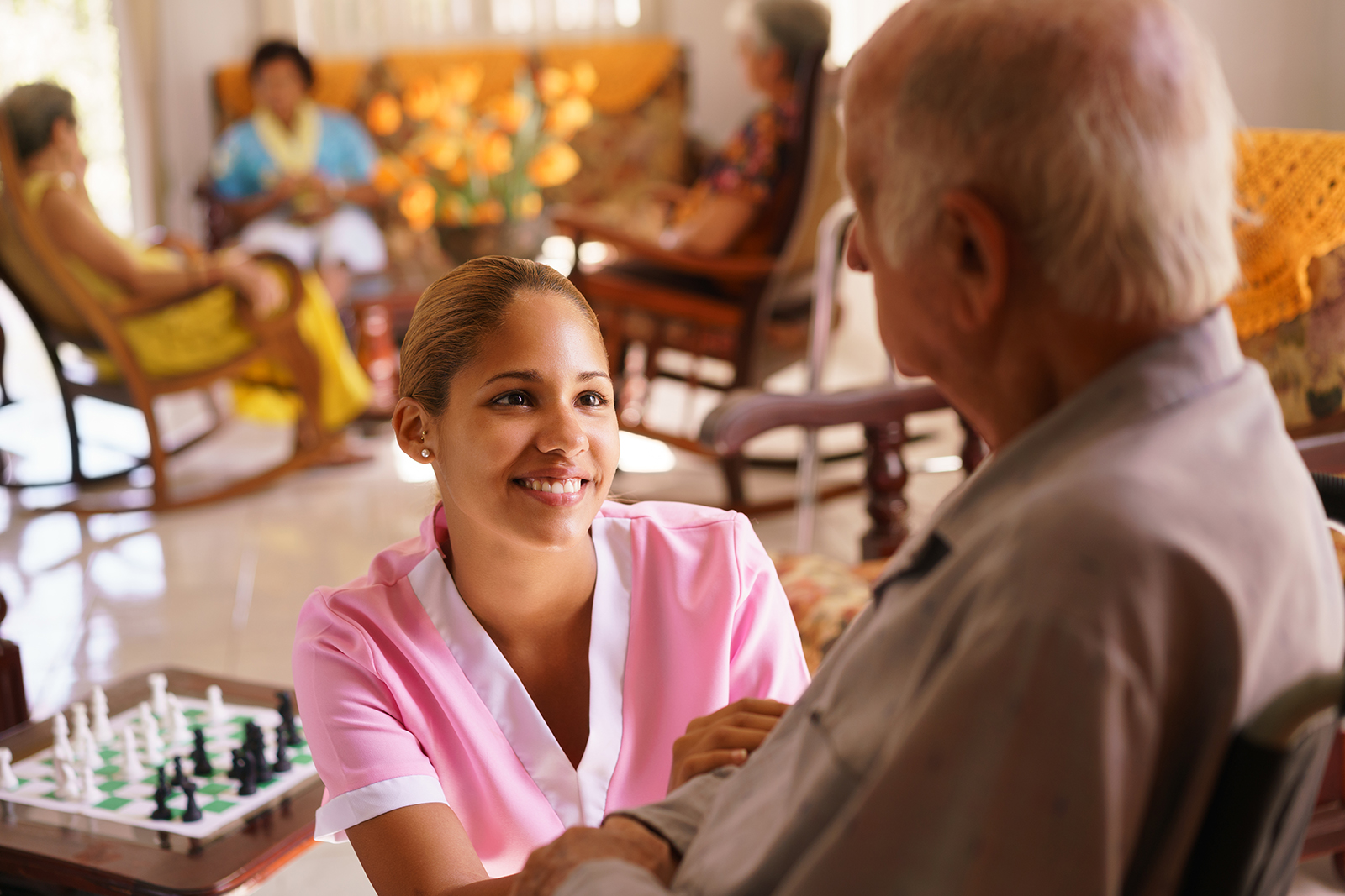 A young nurse is kneeling and smiling in front of a seated elderly man in a nursing home