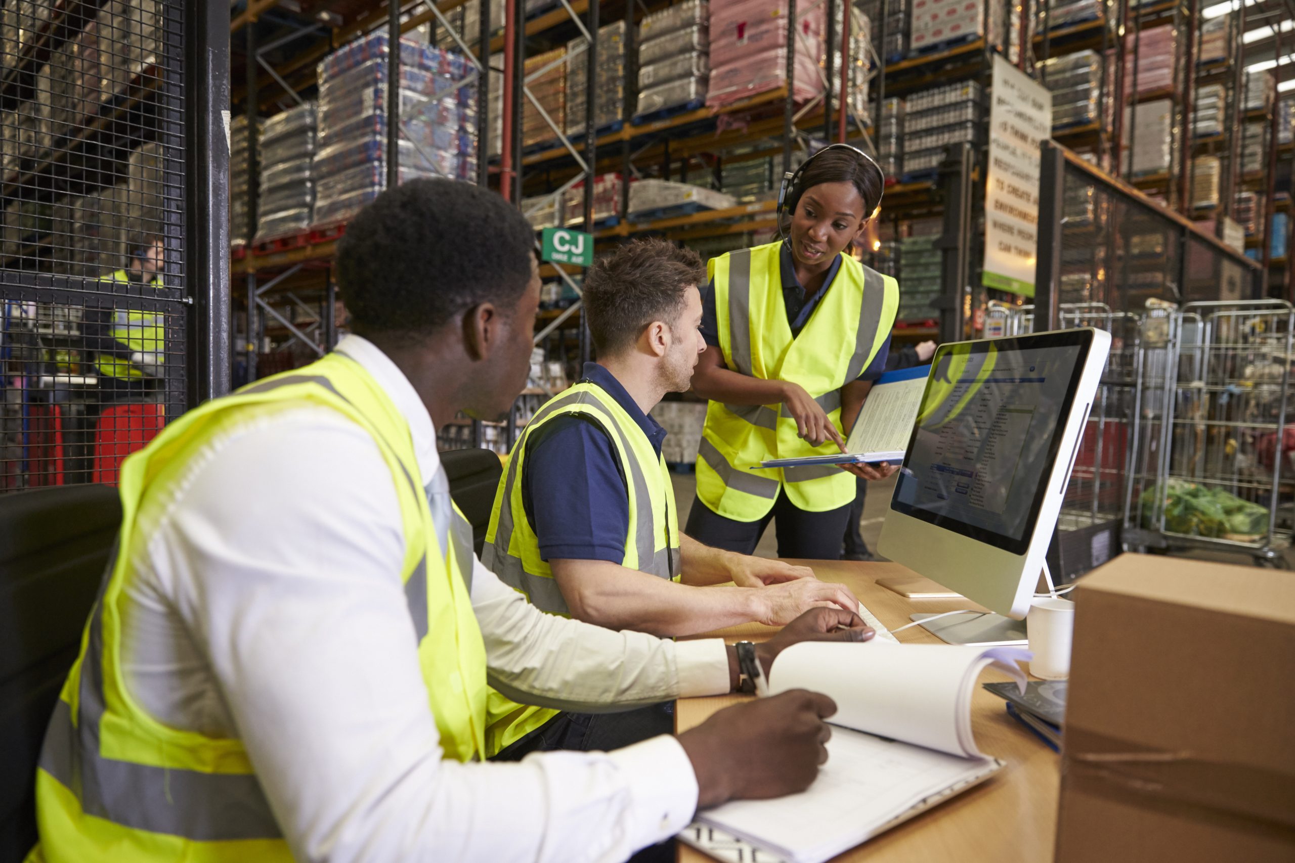 Three workers in yellow vests in a warehouse reviewing paperwork