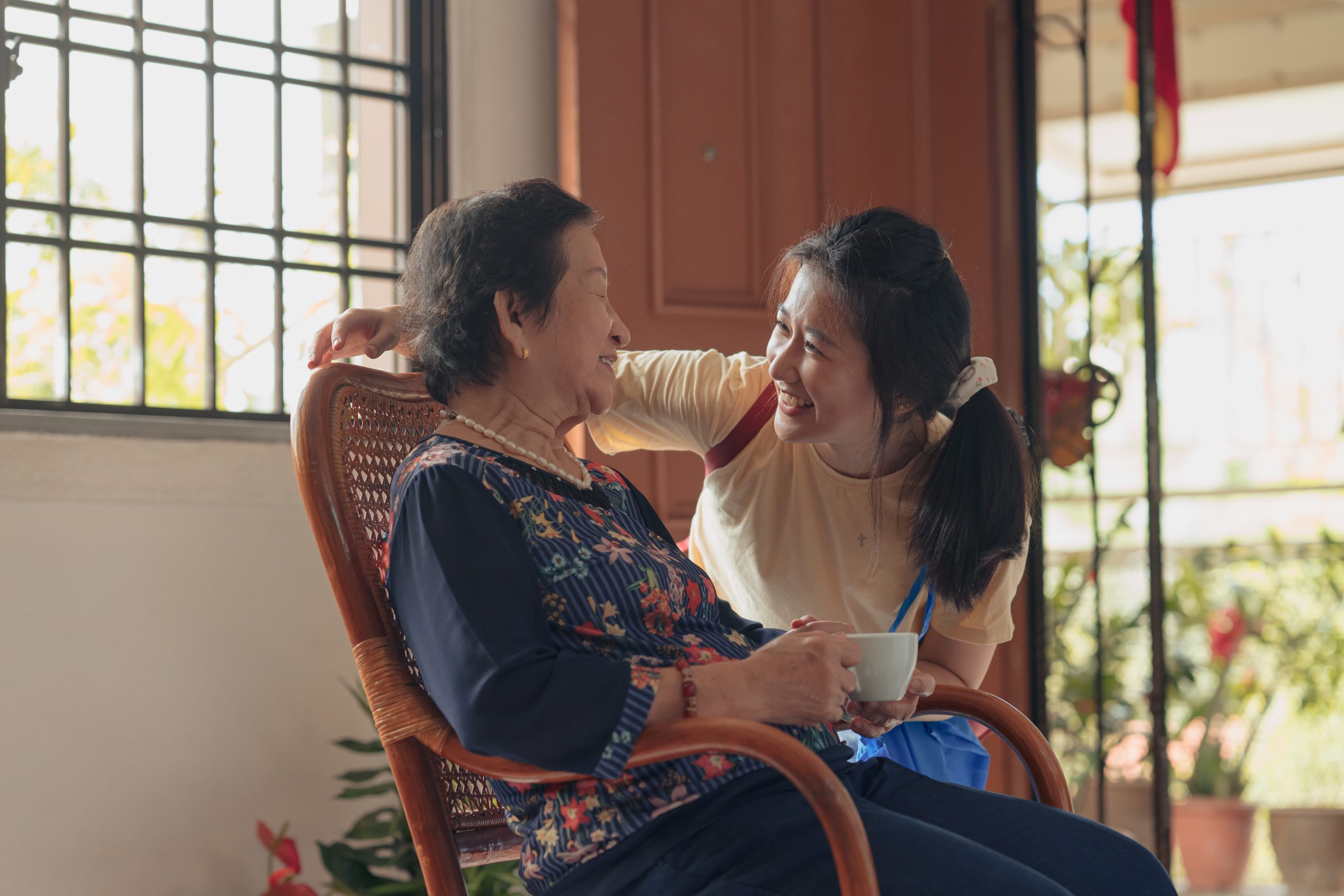 A grandmother is sitting in a rocking chair with coffee. Her granddaughter is smiling and putting her arm around her.