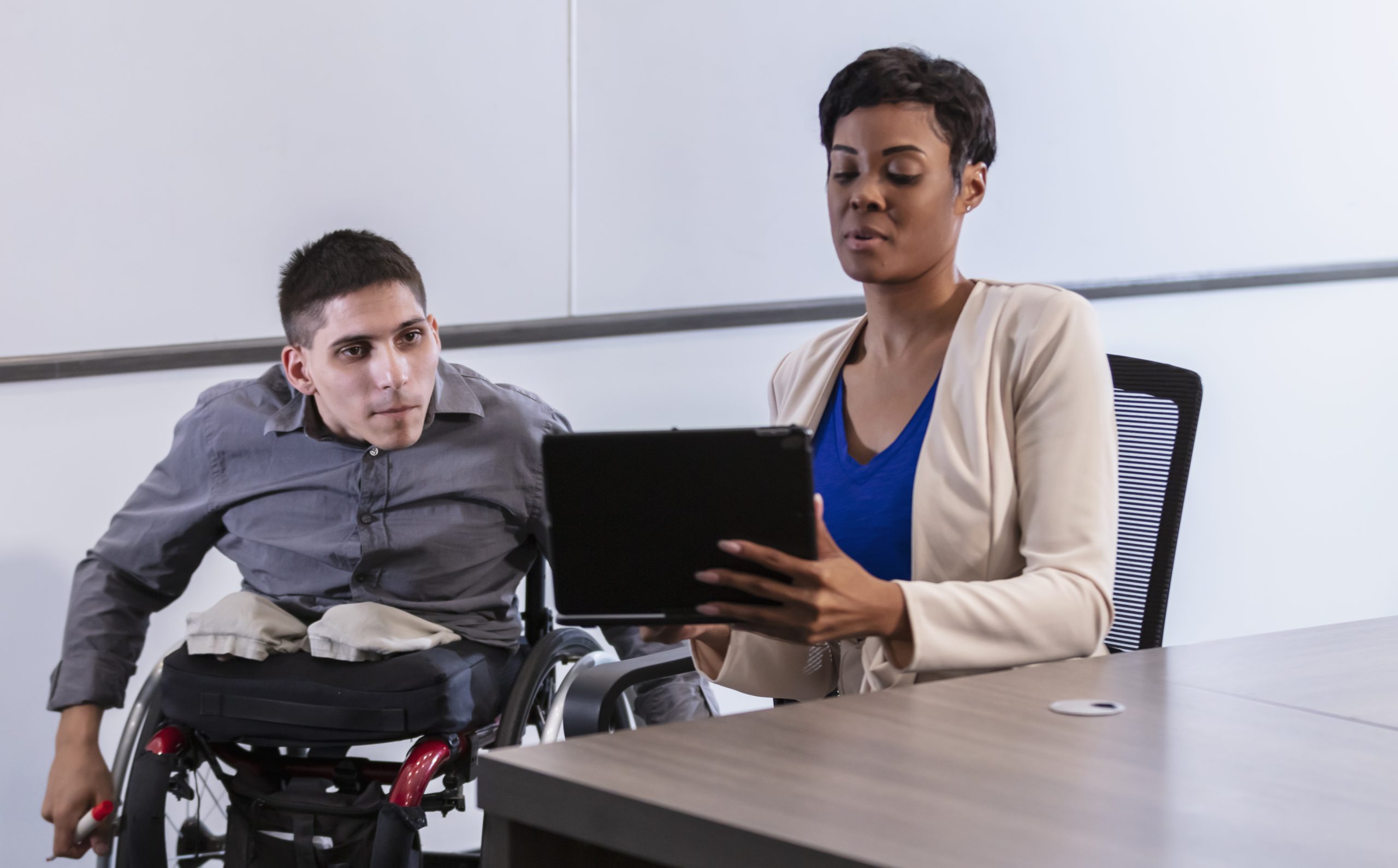 A man in a wheelchair talks with a coworker who is looking at her laptop