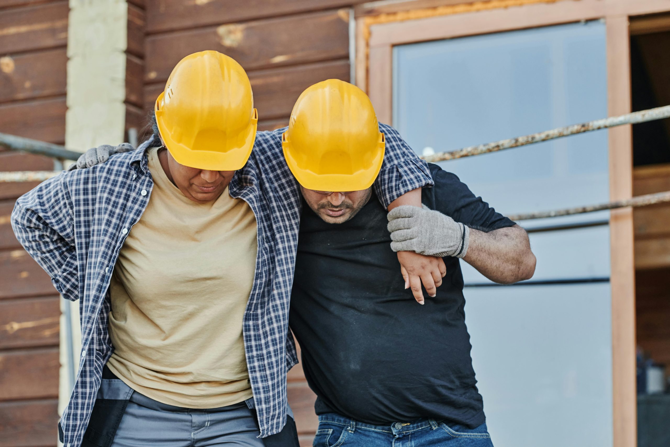 Two men in hardhats, one is injured and needs the support of the other as they walk