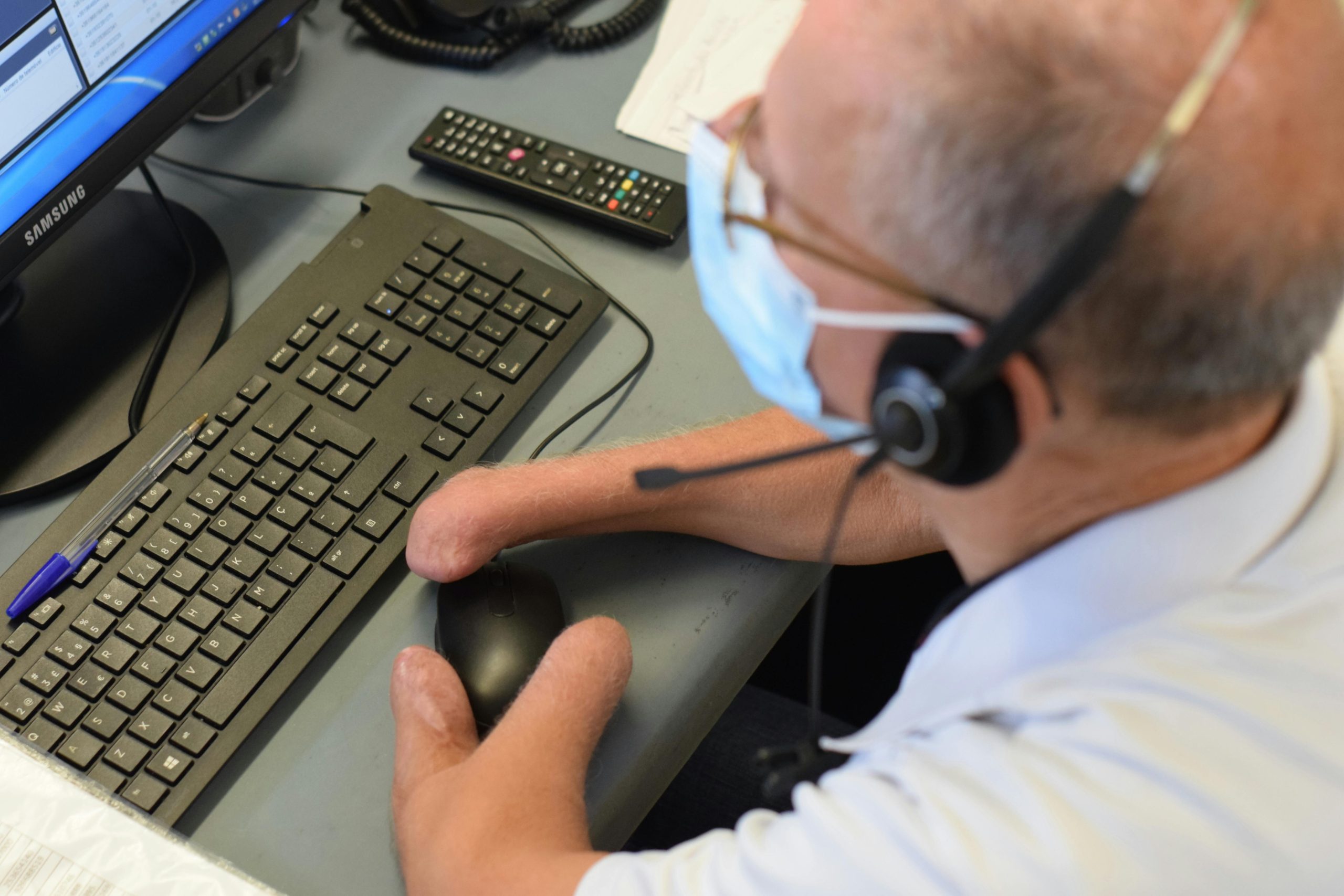 Man wearing mask and a headset at a computer workstation. He doesn't have hands, so he is using an adaptive mouse.
