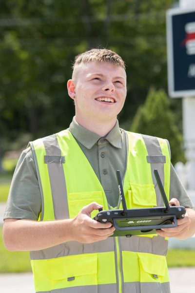 A young man wearing a safety vest uses a drone remote control while looking towards the sky