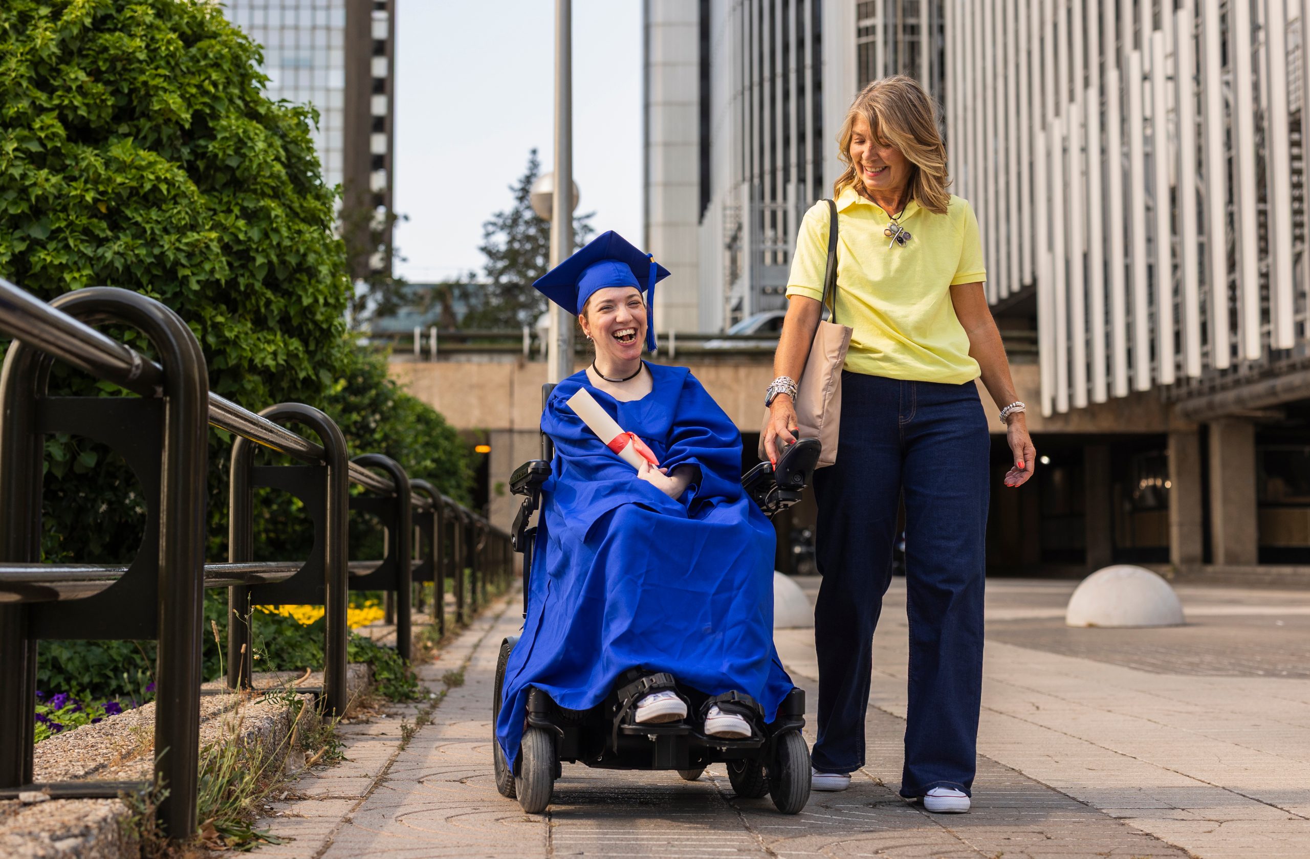 A laughing student in a wheelchair holds a diploma. Her proud mother walks beside her.