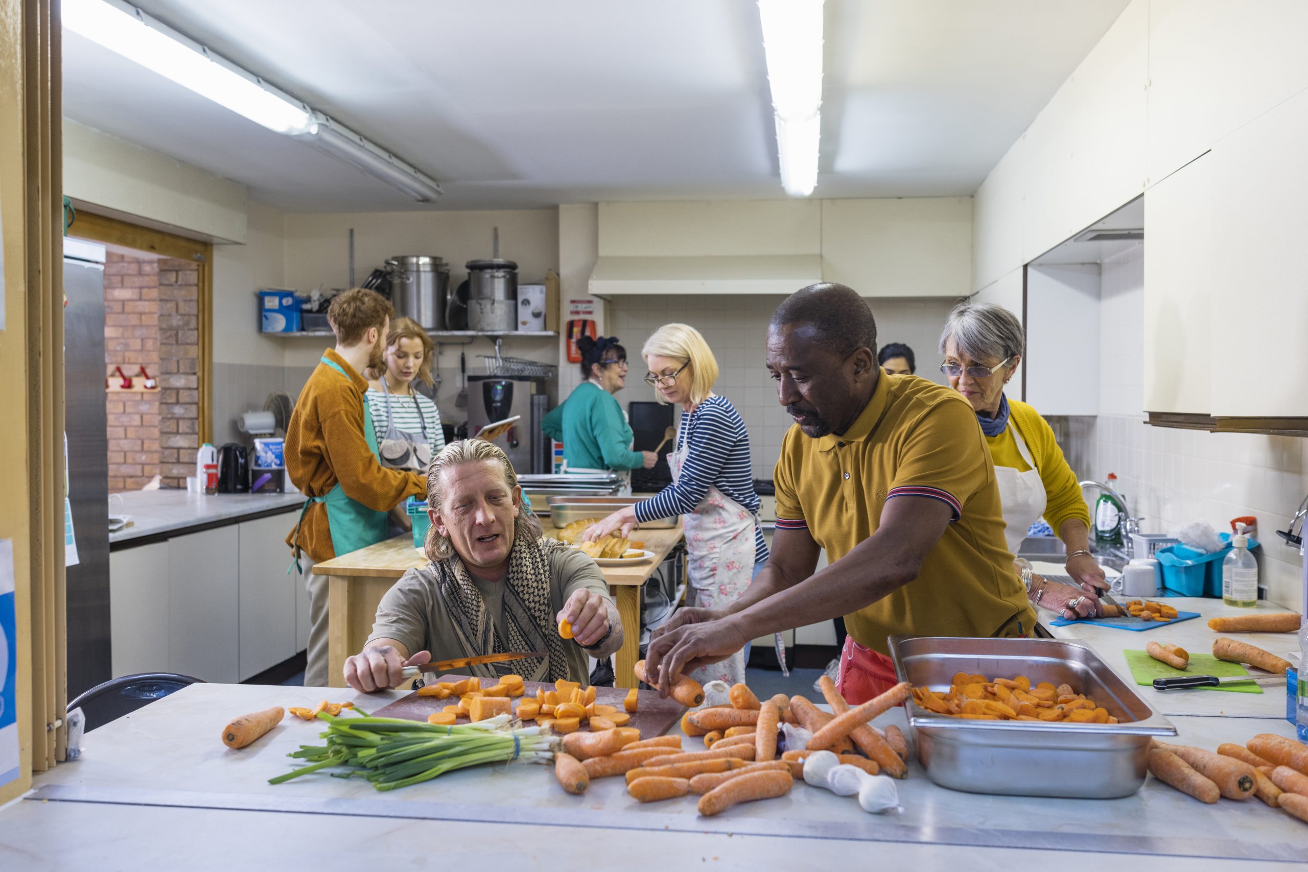 A diverse group of people of all ages, races and abilities chopping vegetables and working in an industrial kitchen to prepare a large meal