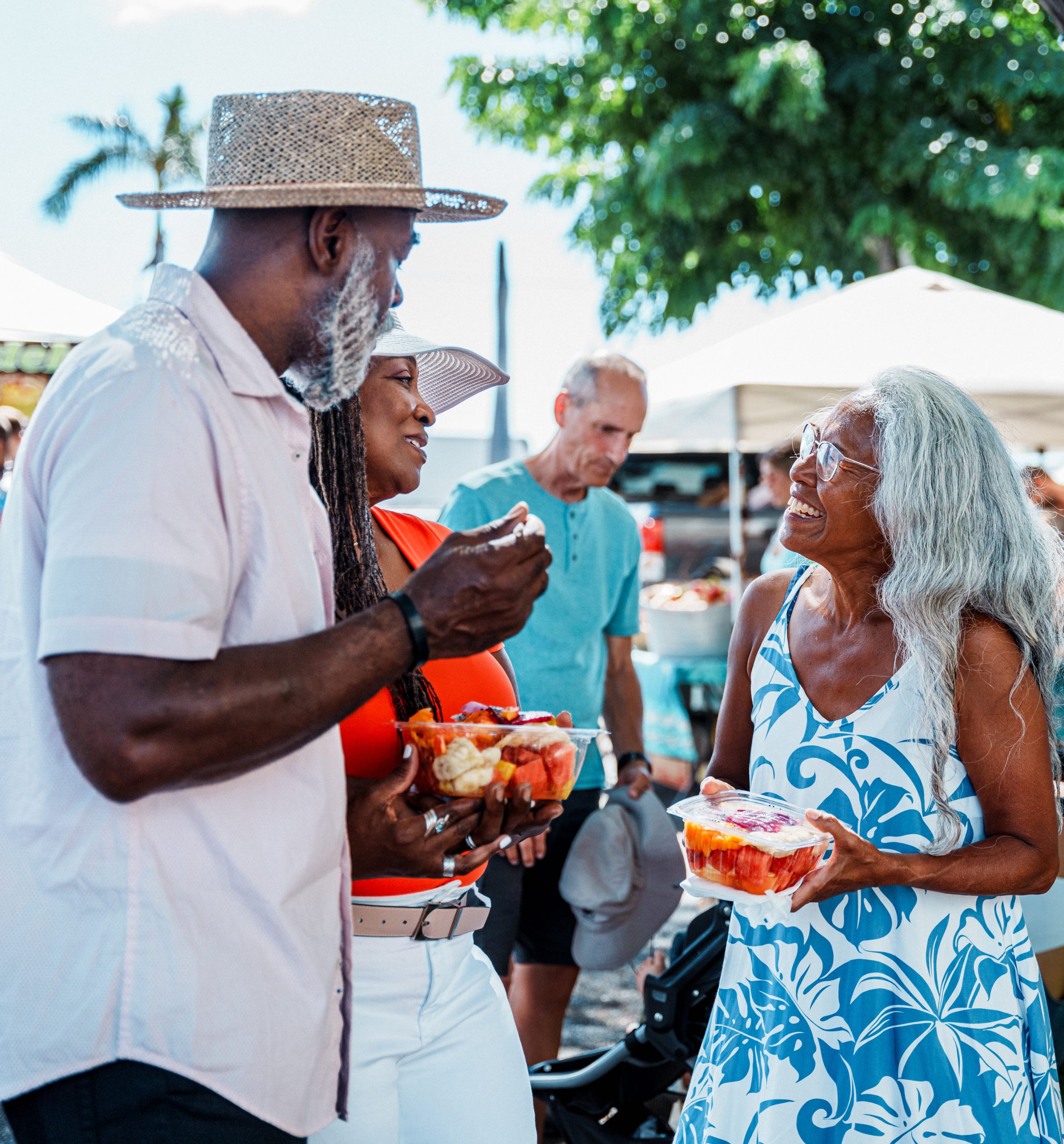 An African American senior couple eat fresh fruit and cheerfully chat with their local tour guide, a senior woman of Hawaiian decent, while at a farmer's market
