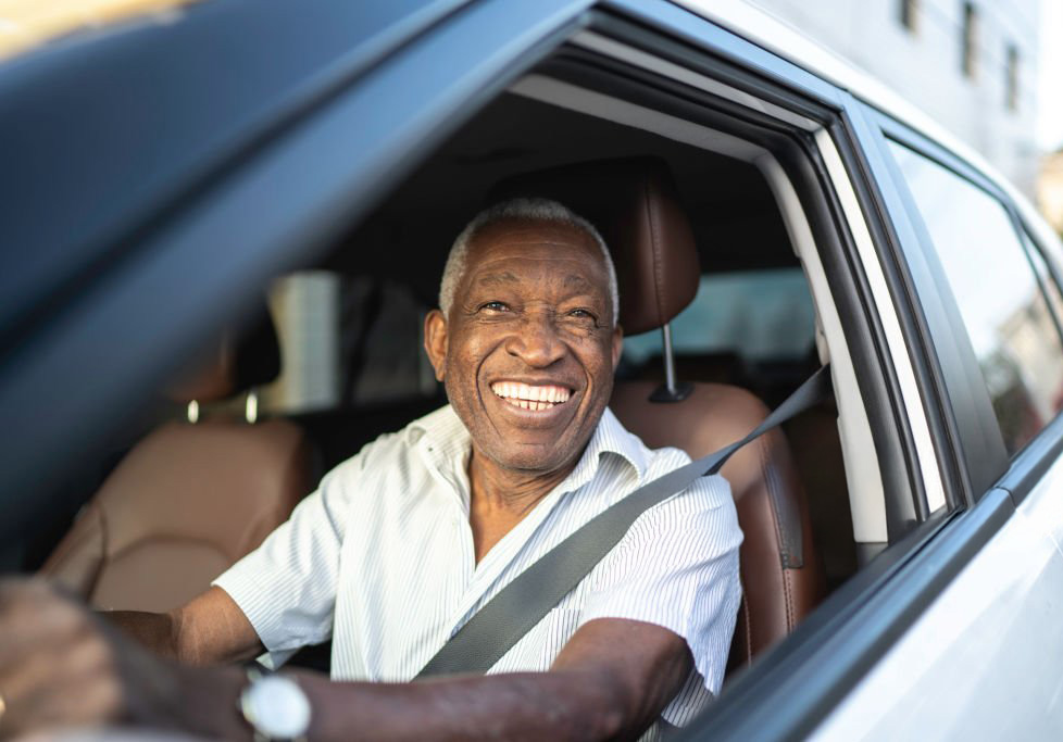 Older man drives a car. He is smiling with hands on the wheel.